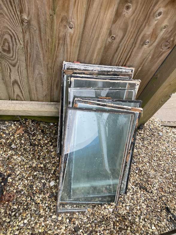 A stack of old, rectangular window panes with chipped paint, ideal for draught proofing projects, leaning against a wooden fence on a gravel surface.