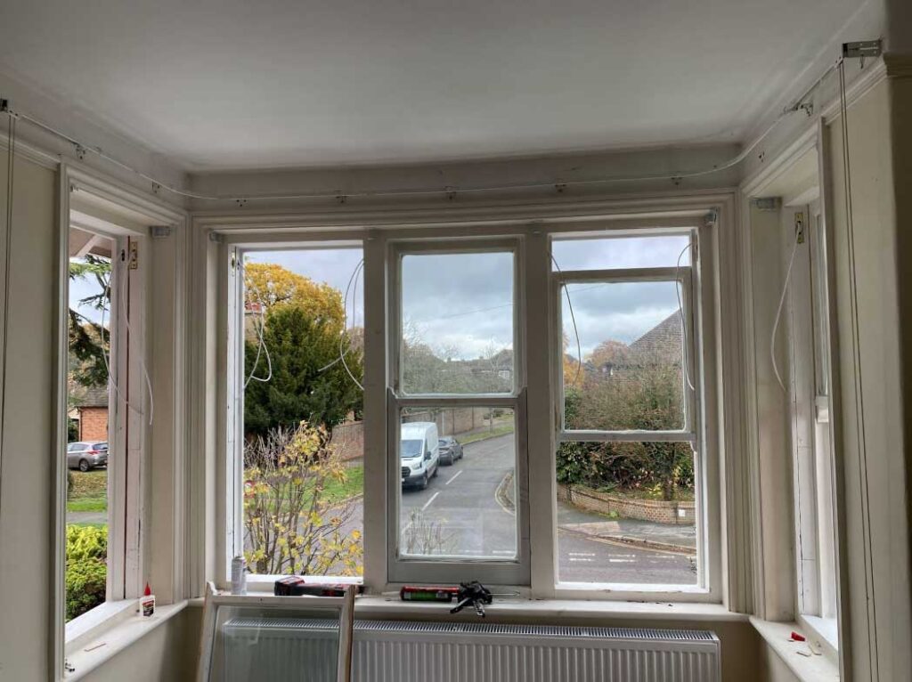 A view from inside a room looking out through three-part bay windows. The window panes are partially open, revealing a suburban street and foliage outside, with draught proofing ensuring comfort within.