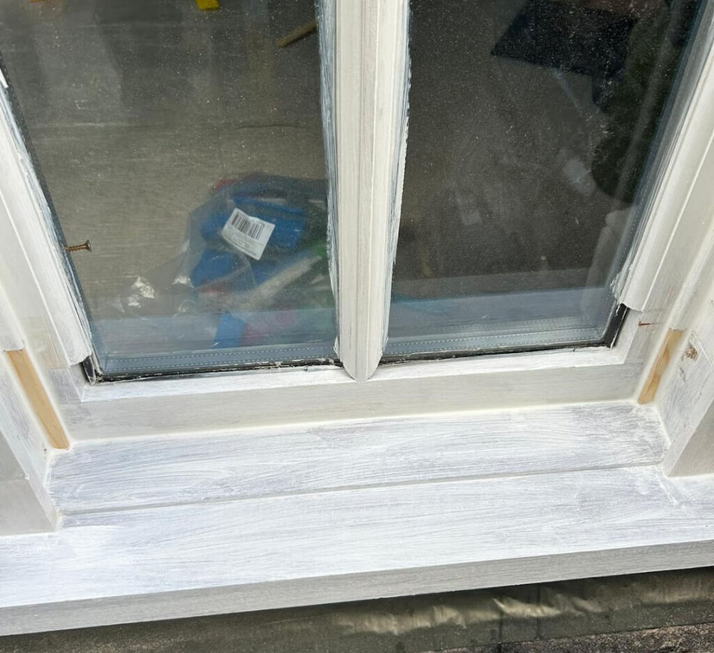 A close-up of a double-paned window with white paint applied to the frame and sill, showcasing the fine results of Sash Windows Repairs Croydon. Various objects are visible through the glass.