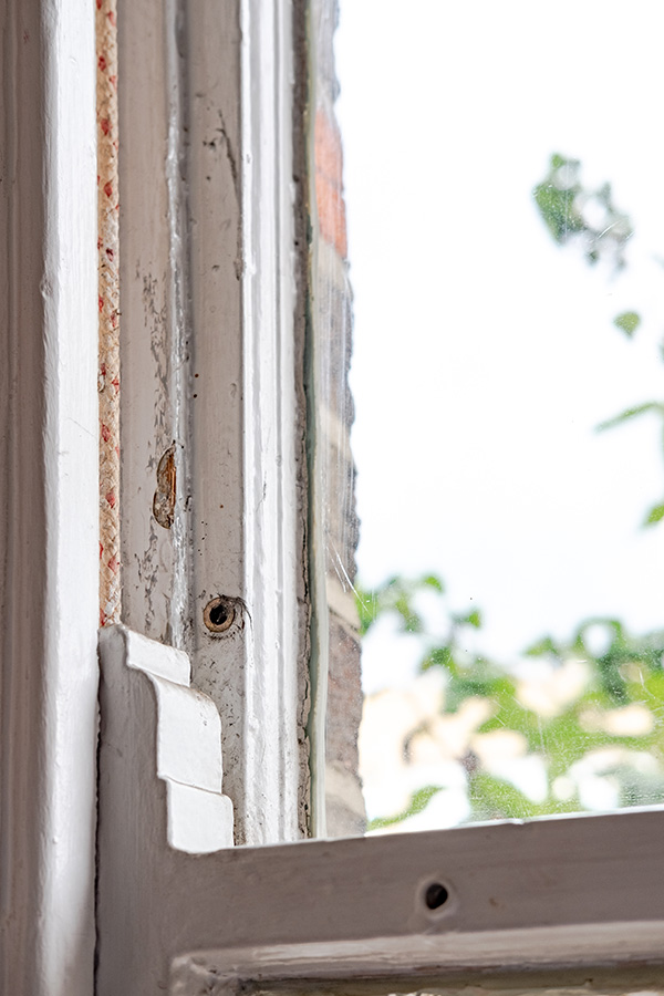 Close-up view of an open wooden window frame with chipped paint, revealing an outdoor scene with blurred greenery in the background, ready to be replaced by new double-glazed sashes.