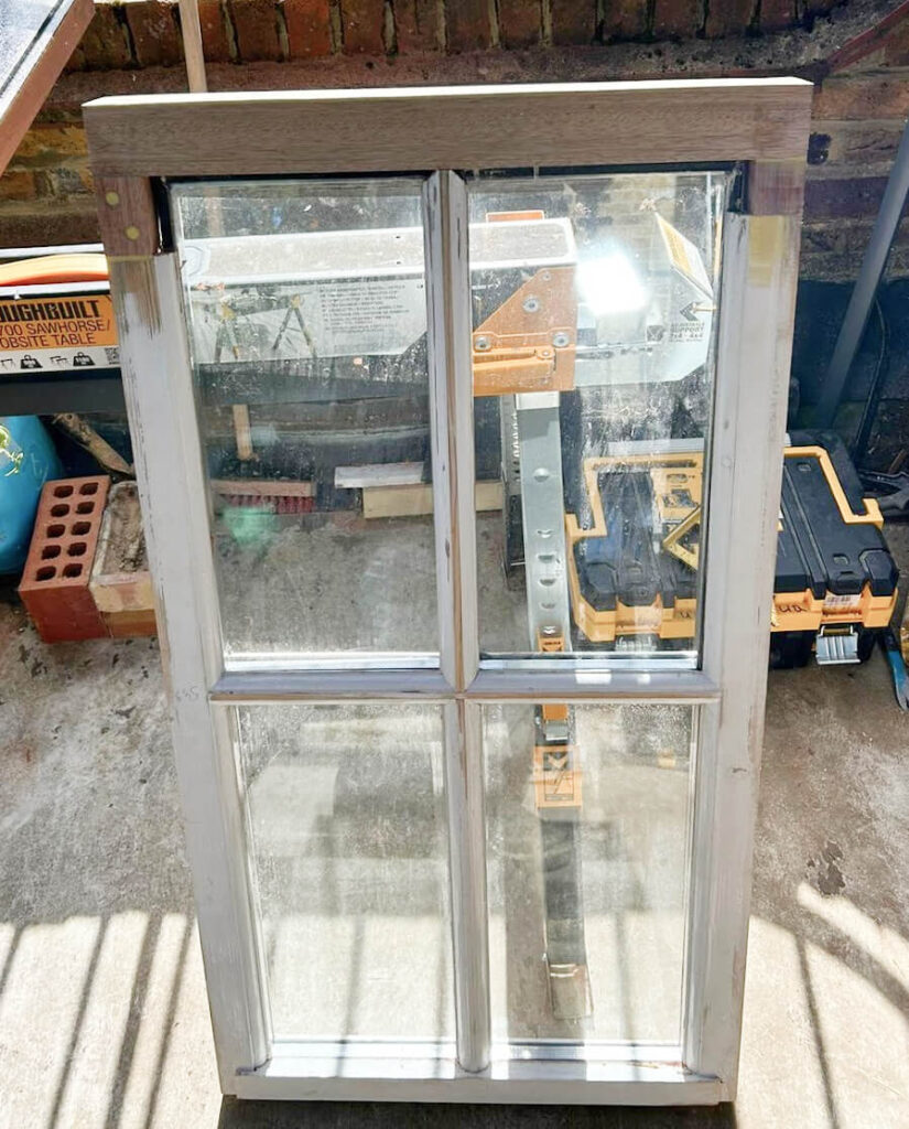 A four-pane glass window with a white frame, awaiting Sash Windows Repairs Croydon, is propped up against a brick wall in a workshop, surrounded by various tools and construction materials.