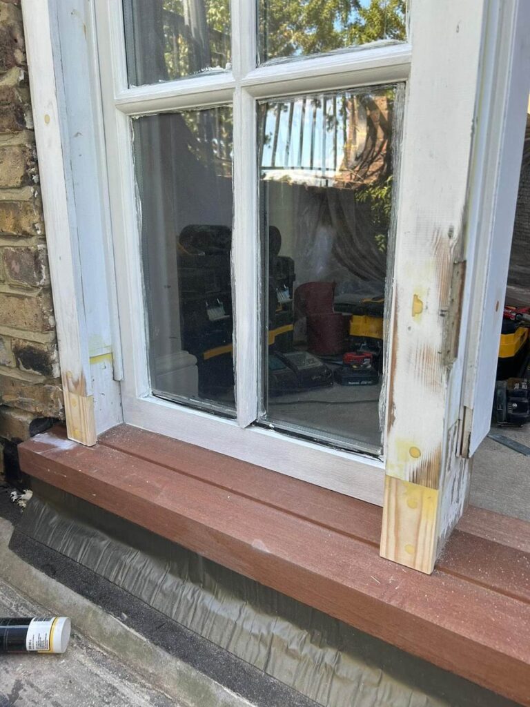 Window frame and window sill repair underway with wood filler. Some areas are sanded and ready for painting. Tools and equipment are visible inside through the glass.