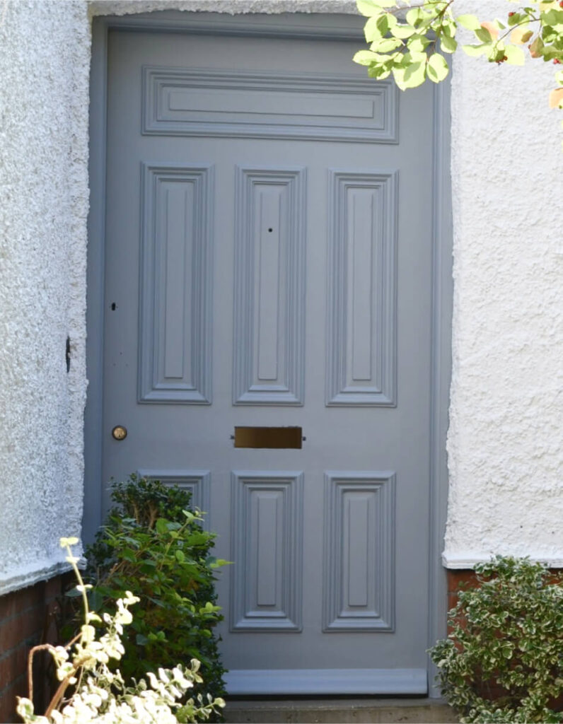 A closed grey-paneled door, recently enhanced through door restoration, features a brass mail slot and knob, elegantly framed by white walls and vibrant green shrubs.