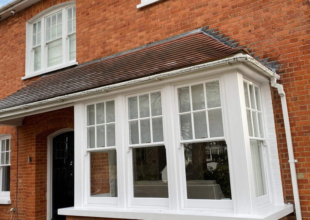 A charming red brick house featuring a white bay window with sash spiral balance, and a black front door. The window boasts a tiled roof and white frames, embodying classic elegance.