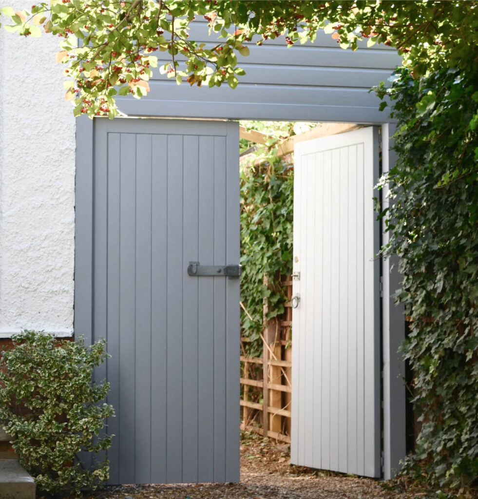 An open wooden gate, recently restored with expert door restoration techniques, leads to a garden path surrounded by lush greenery.