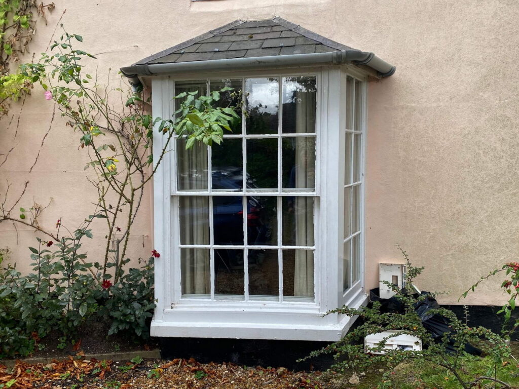 A house with a small bay window featuring sash spiral balance repair overlooks lush greenery, including a rose bush with a few red flowers.