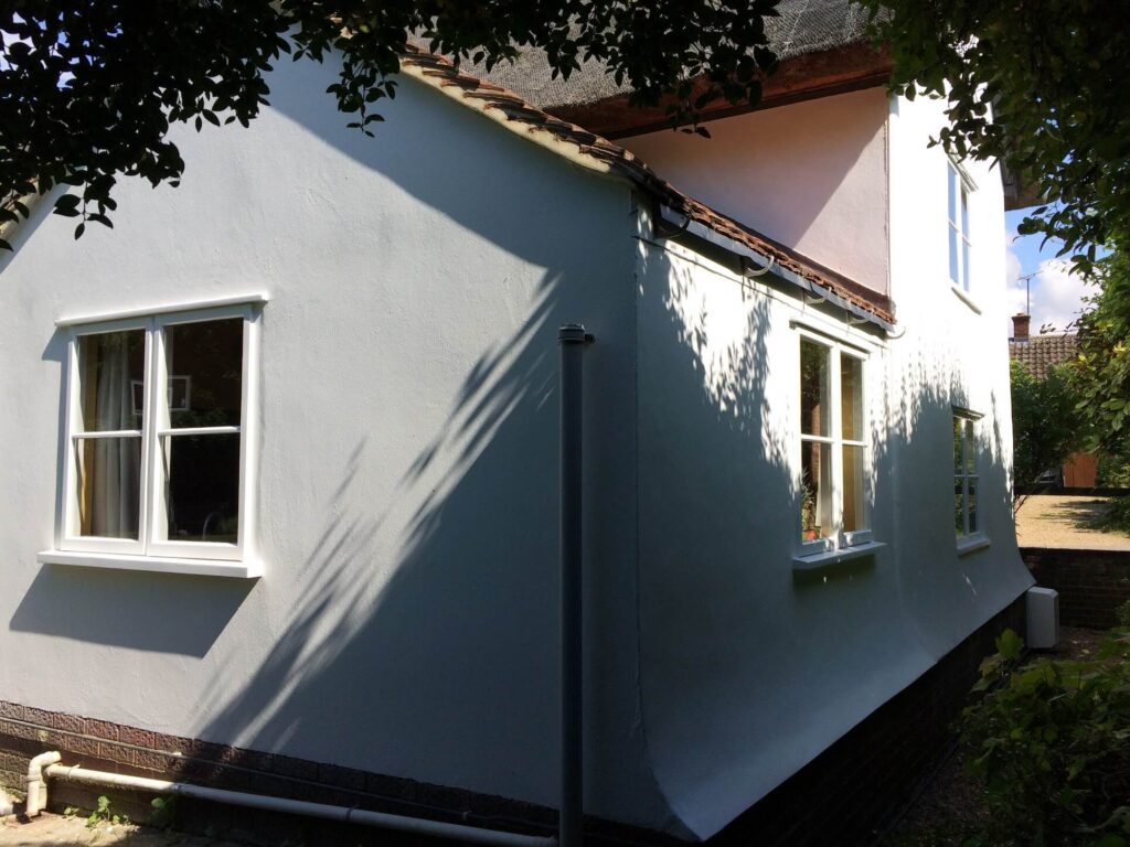 A white house with a sloped roof and wooden window painting is partially shaded by trees, enhancing its charming rustic appeal.