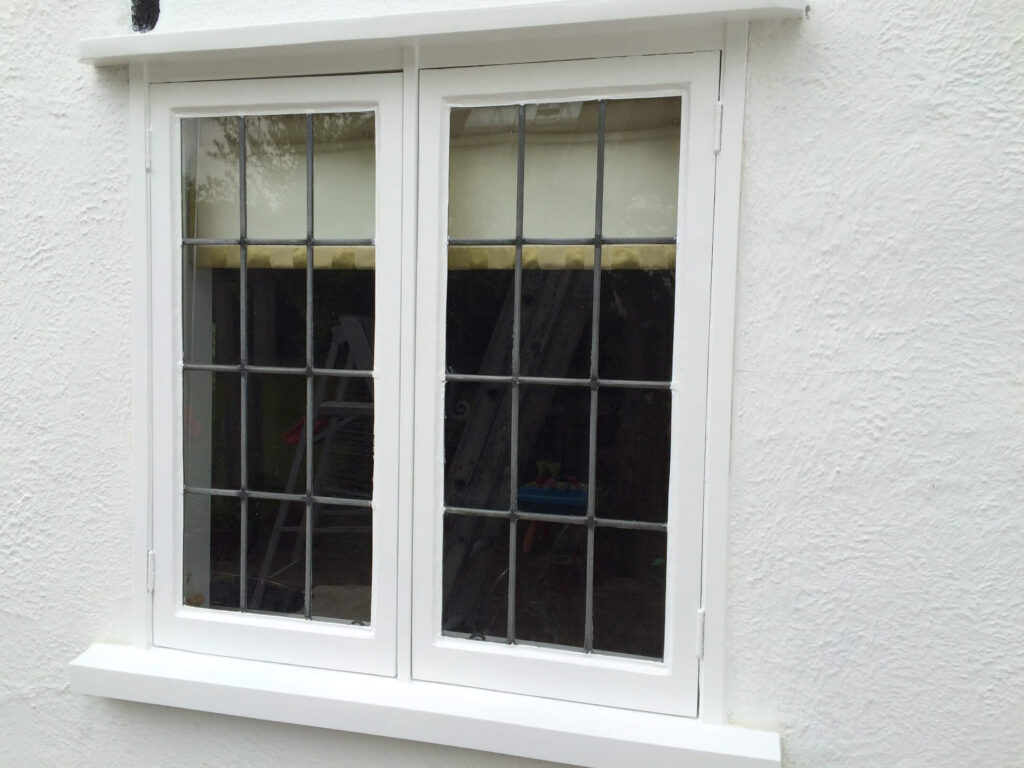 A beautifully crafted white-framed window, reminiscent of a wooden window painting, features grid-patterned glass panes set perfectly against a textured, white wall.