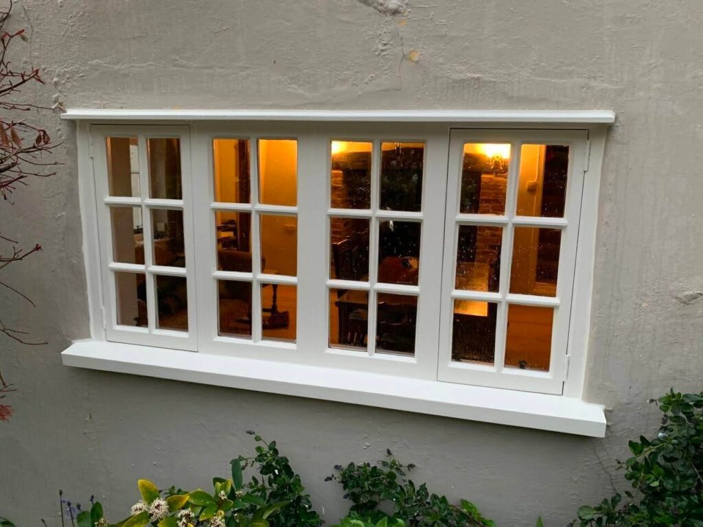 A wooden multi-pane window with warm light glowing from inside, set in a textured wall, surrounded by foliage at the bottom, evokes the charm of a thoughtful restoration.