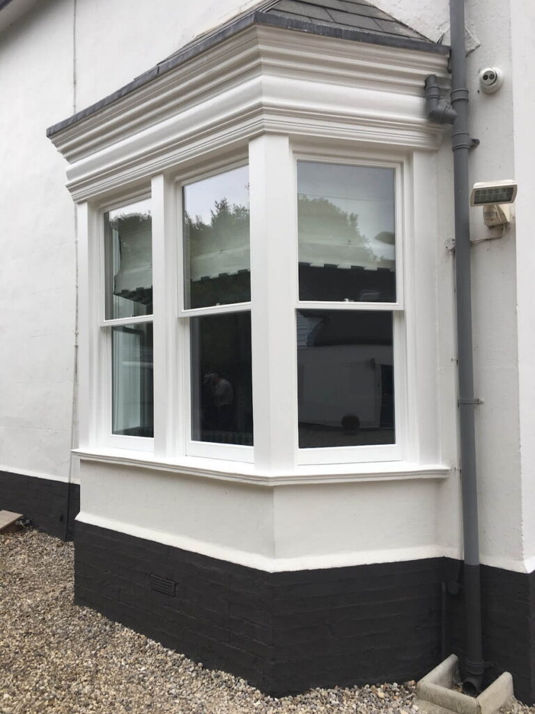 A white bay window with a sash spiral balance in need of repair rests on a black base trim, accentuating the light-colored building surrounded by gravel.