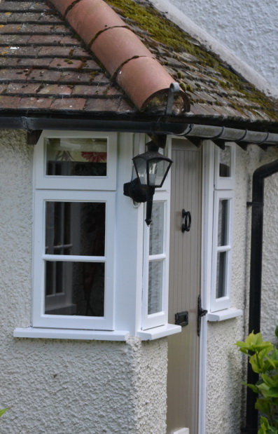 A white cottage exterior with a small porch showcases a hanging lantern and a charming wooden door, beautifully accentuated by restored wooden-framed windows.