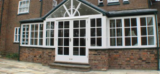 A charming brick house features a large white-framed glass sunroom extension with double doors, enhanced by a recent wooden window restoration that adds timeless elegance.