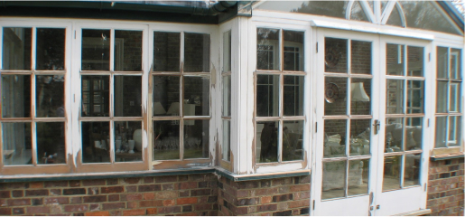 A section of a building boasts wooden windows in white frames, their paint chipped to reveal the original texture beneath. Large glass panes and a brick wall form the lower half, inviting thoughts of restoration to preserve its timeless charm.