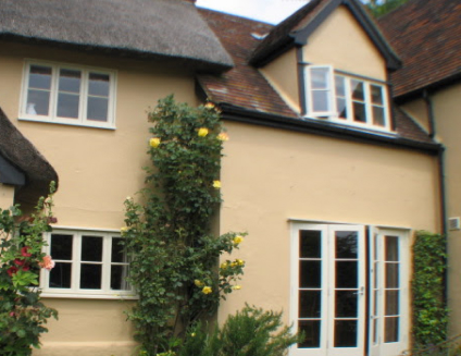 A beige two-story house with white and wooden-framed windows, a thatched roof, and a charming wooden door, surrounded by greenery and yellow flowers. Perfectly blending tradition with nature's embrace through careful restoration.